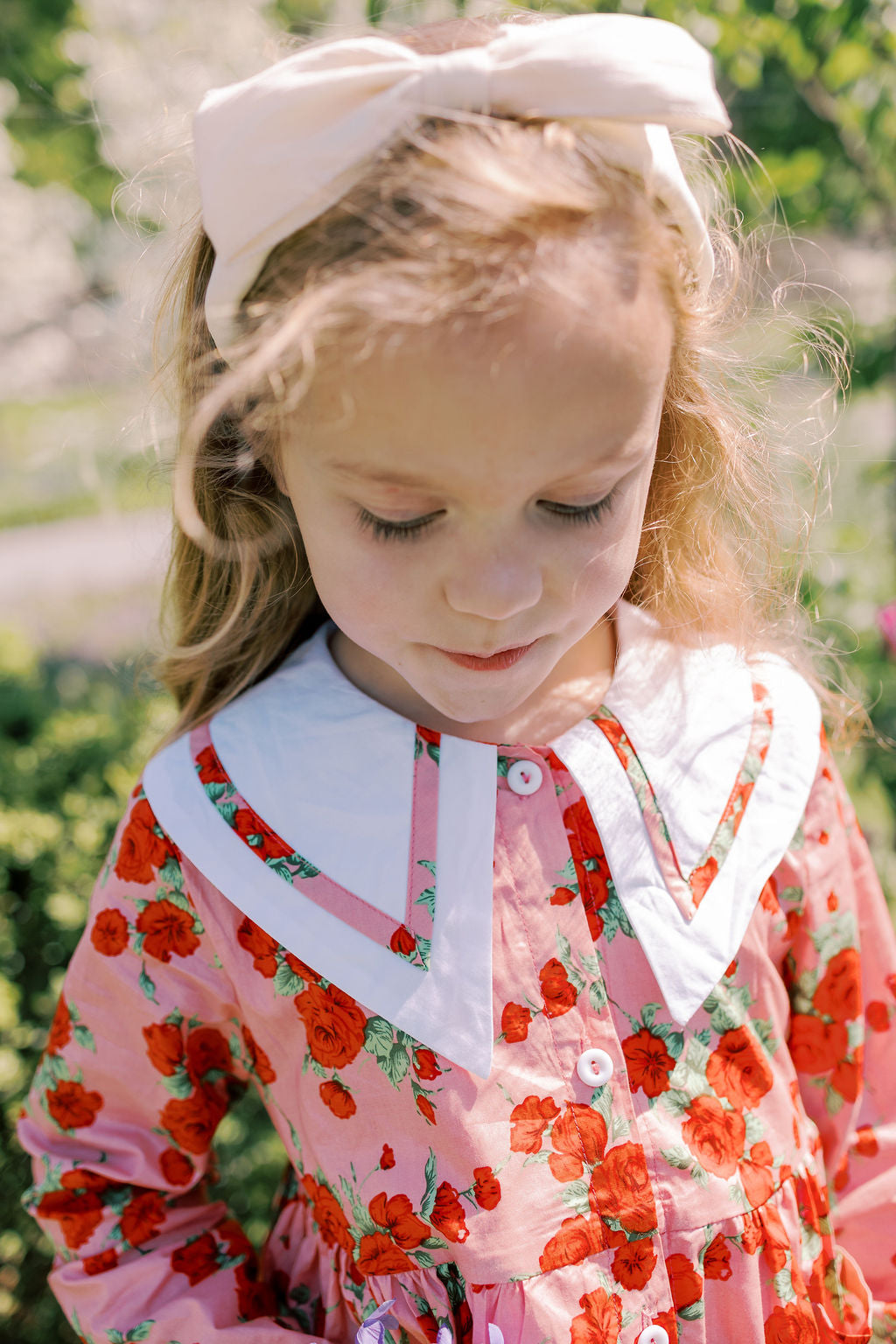 Young girl wearing a floral dress with a large bow in her hair, standing outdoors.