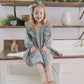 A young girl with blonde hair sits barefoot on a white wooden table in a bright, styled room. She is wearing a long-sleeve, knee-length dress with a purple and white gingham pattern and a matching bow headband.