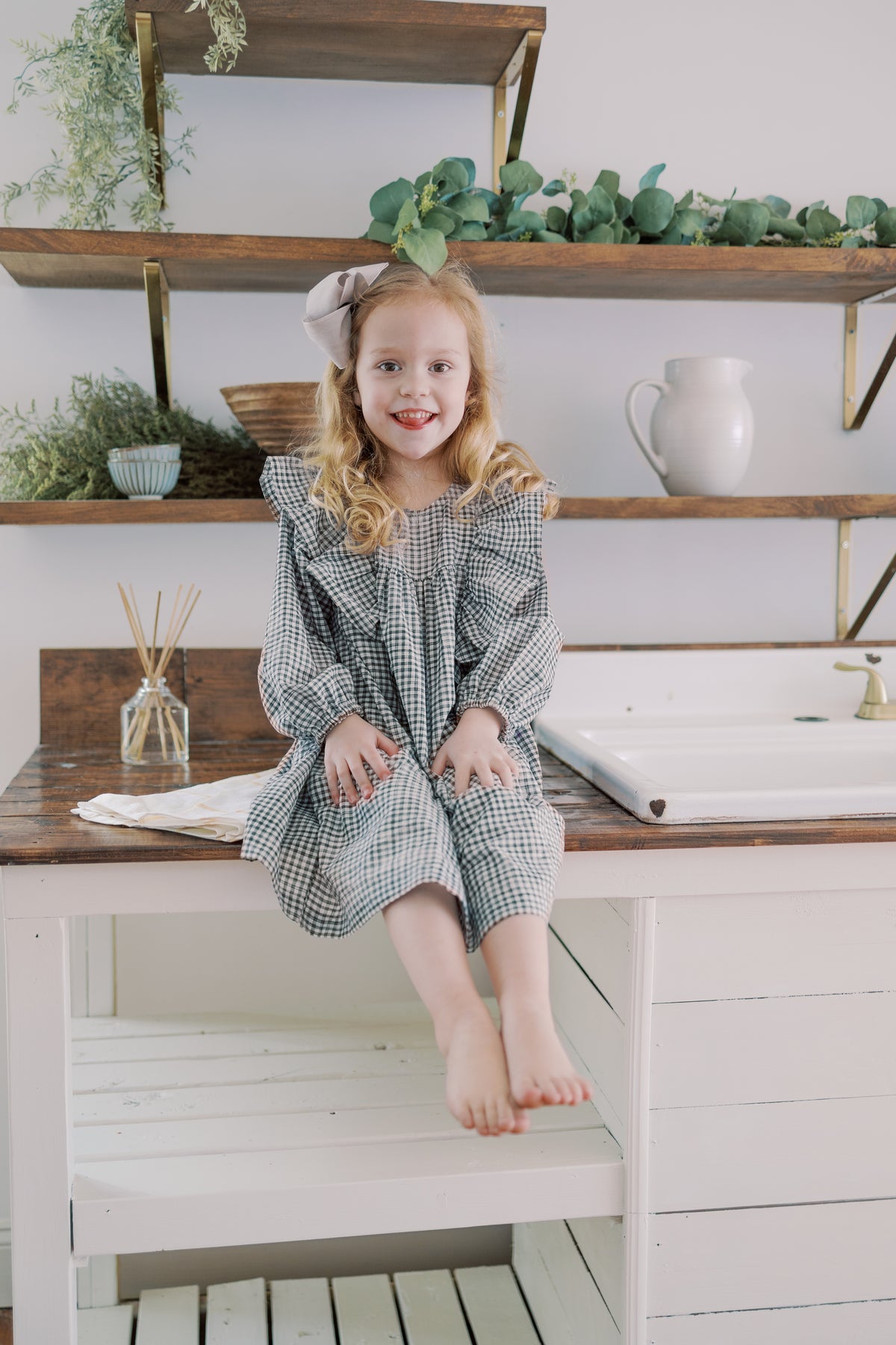 A young girl with blonde hair sits barefoot on a white wooden table in a bright, styled room. She is wearing a long-sleeve, knee-length dress with a purple and white gingham pattern and a matching bow headband.