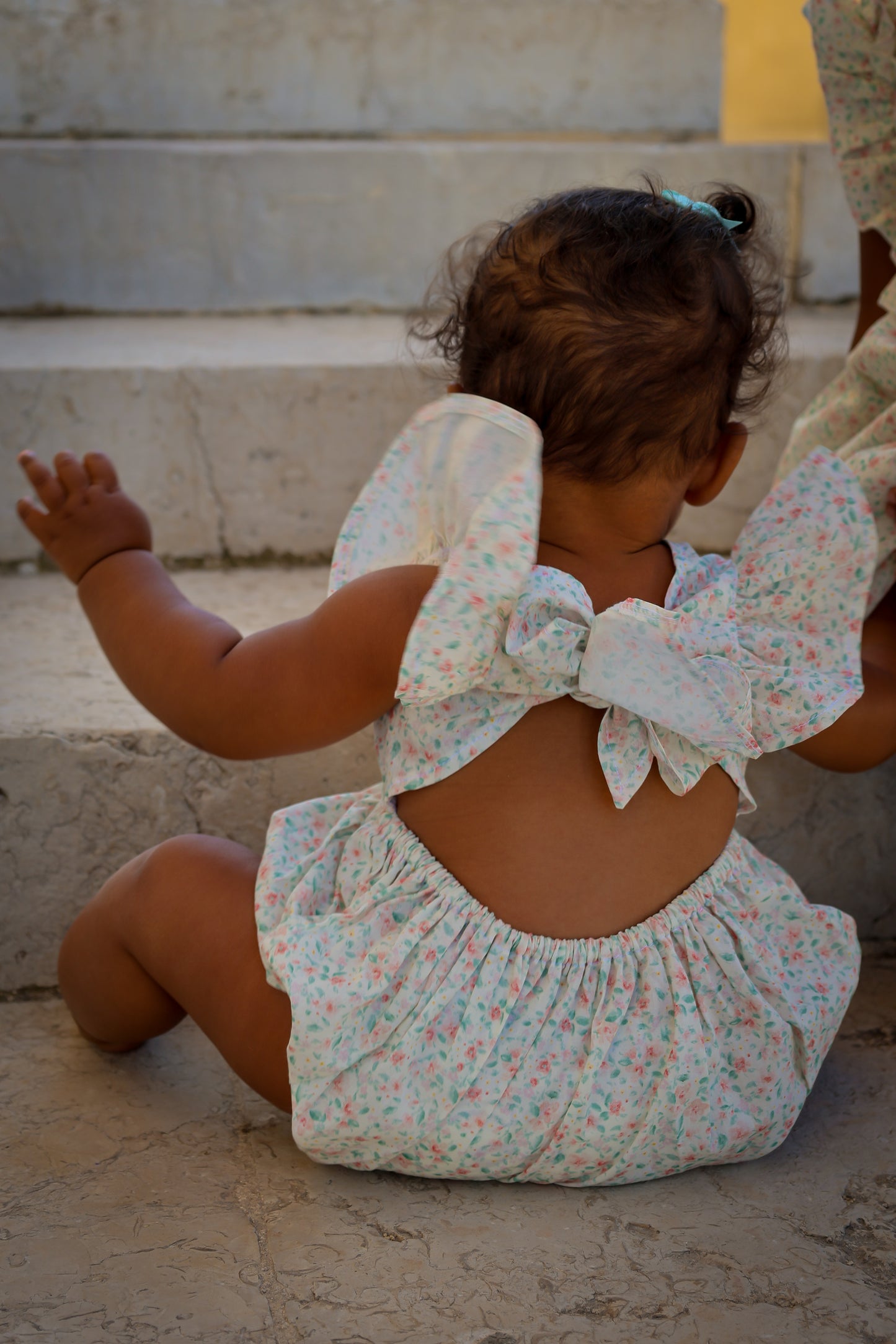 Baby in a floral outfit with a large bow sitting on stone steps.