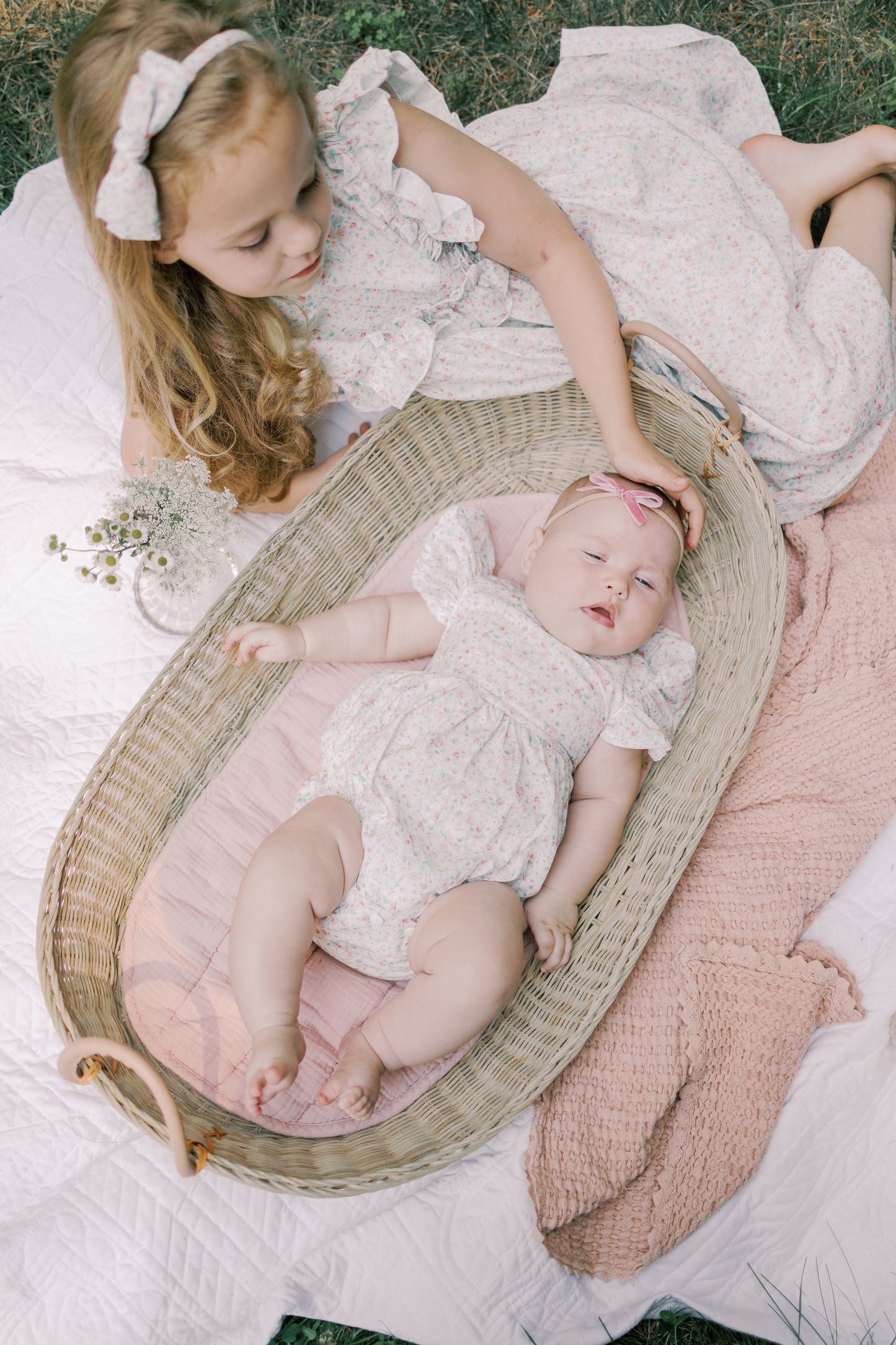 Two children in matching outfits lying on a blanket with a baby in a Moses basket.