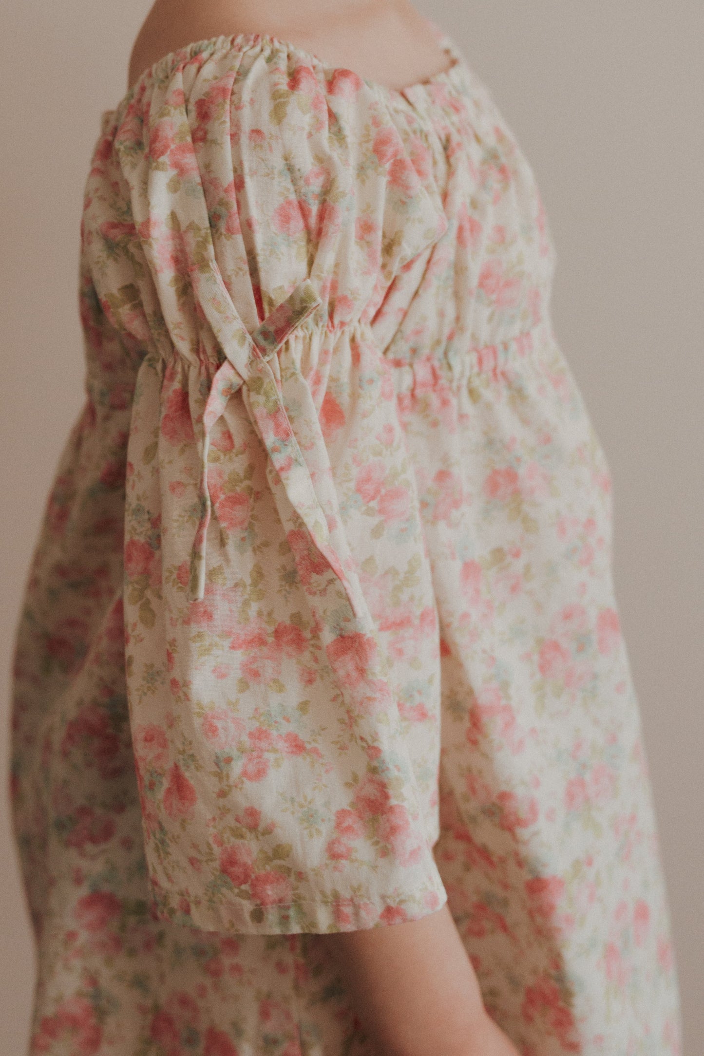 A young girl wearing a pink floral cotton dress with bell sleeves, holding a pink flower, with her hair styled in a bun.
