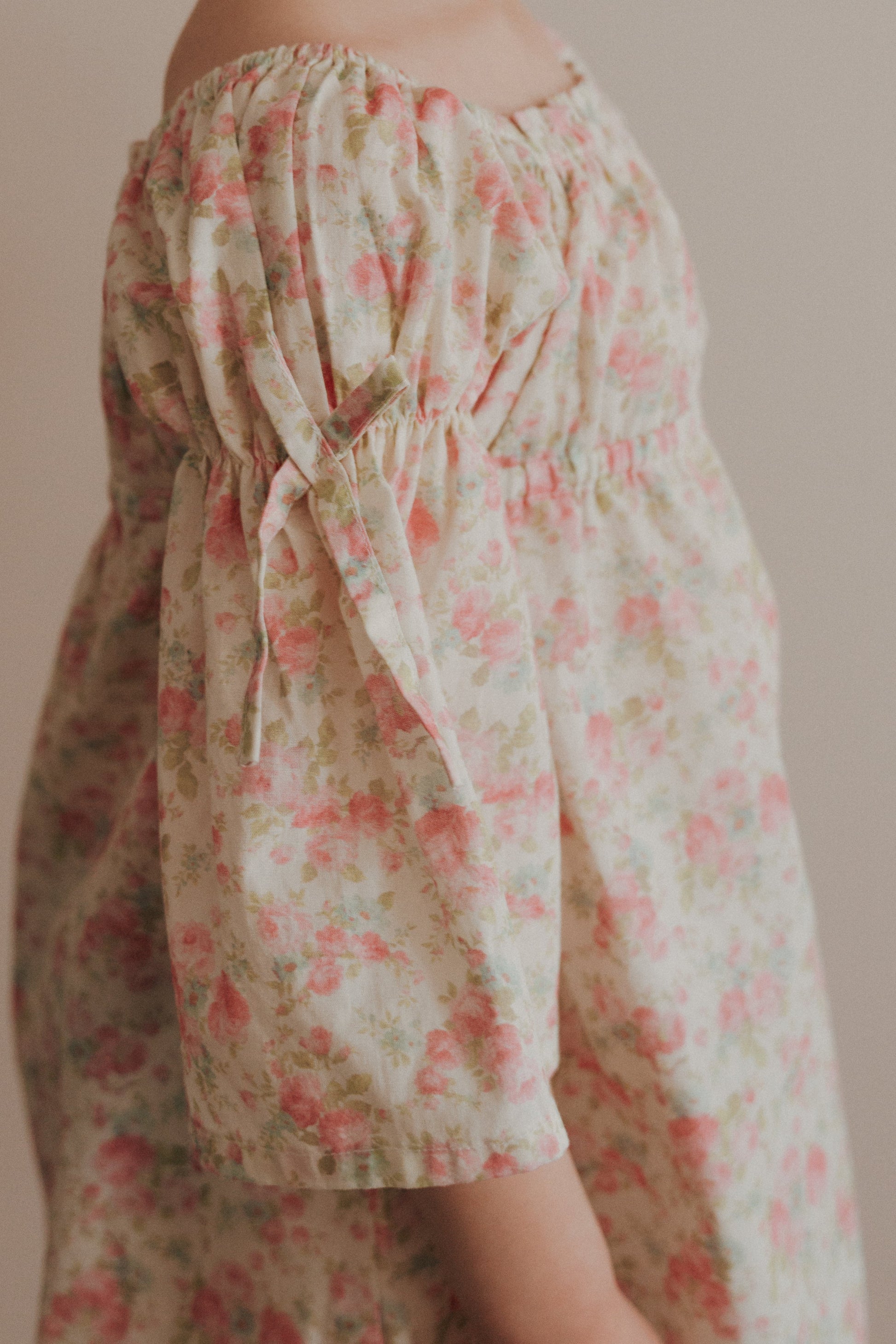 A young girl wearing a pink floral cotton dress with bell sleeves, holding a pink flower, with her hair styled in a bun.