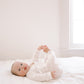 A baby lying on a white bed in a bright, minimalist room, wearing a white long-sleeve romper.