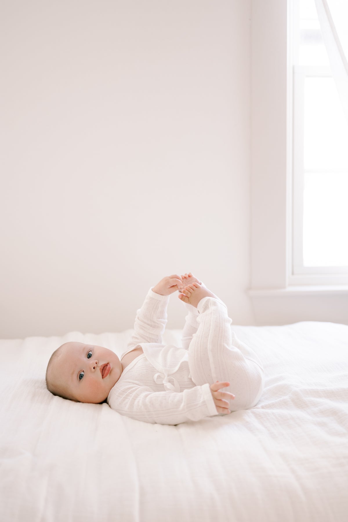 A baby lying on a white bed in a bright, minimalist room, wearing a white long-sleeve romper.