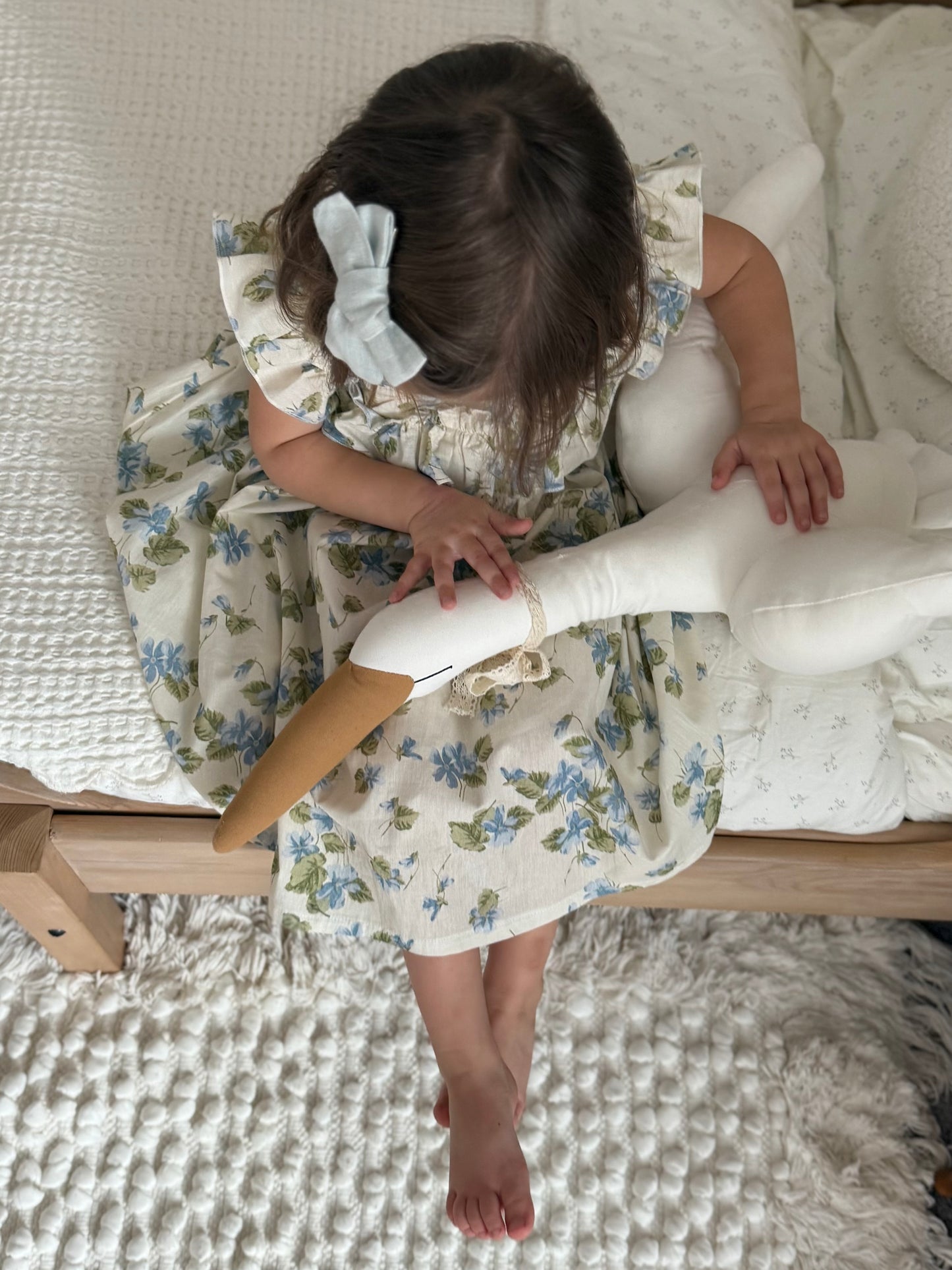 Child in a floral dress sitting on a bed with a wooden hanger.