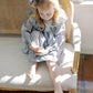 A young girl with blonde hair sits on a vintage cane and wood bench, wearing a purple and white gingham dress with and hair bow. The scene is lit by warm, natural sunlight on a dark wood floor.