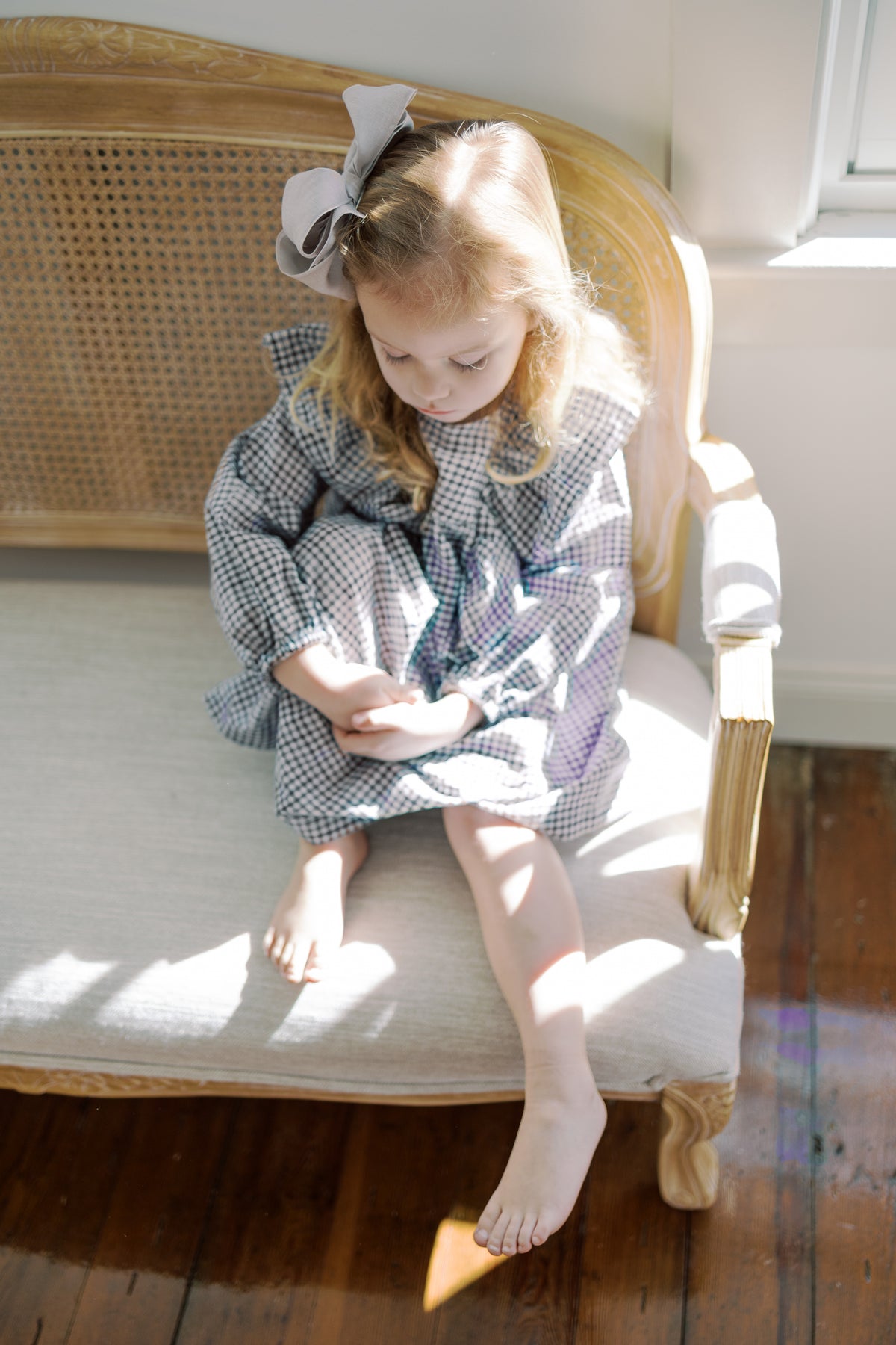 A young girl with blonde hair sits on a vintage cane and wood bench, wearing a purple and white gingham dress with and hair bow. The scene is lit by warm, natural sunlight on a dark wood floor.