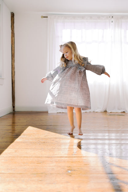 A young girl with blonde hair is barefoot, twirling in a billowy purple and white checkered dress with long sleeves and a ruffled collar, in a bright room with white curtains and a wooden floor.