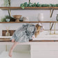 A young girl in a purple and white gingham dress and white bow leans over a white sink in a rustic kitchen setting with wooden shelves and greenery.