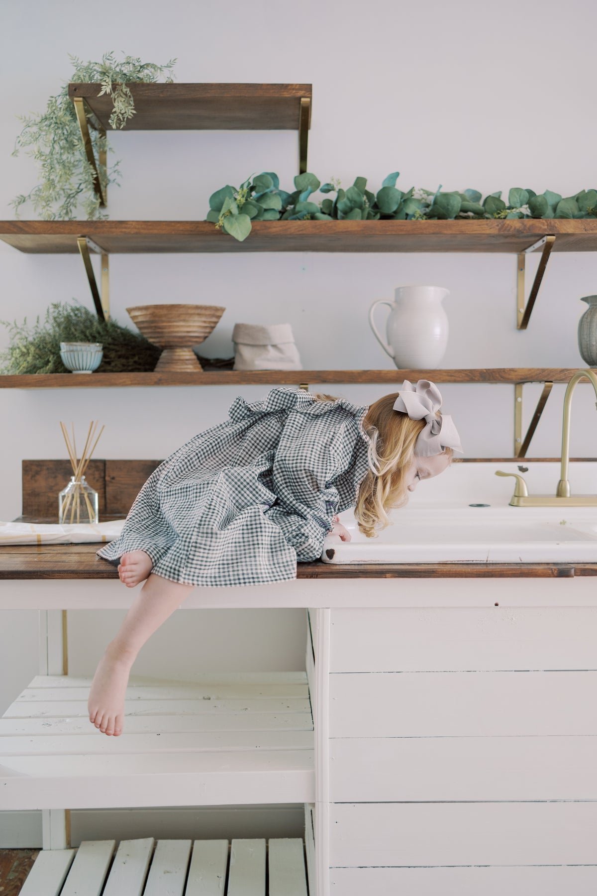 A young girl in a purple and white gingham dress and white bow leans over a white sink in a rustic kitchen setting with wooden shelves and greenery.