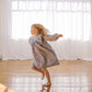 A young girl with blonde hair is barefoot, twirling in a loose, long-sleeved plaid dress or tunic in a bright room with white sheer curtains and a polished wooden floor.