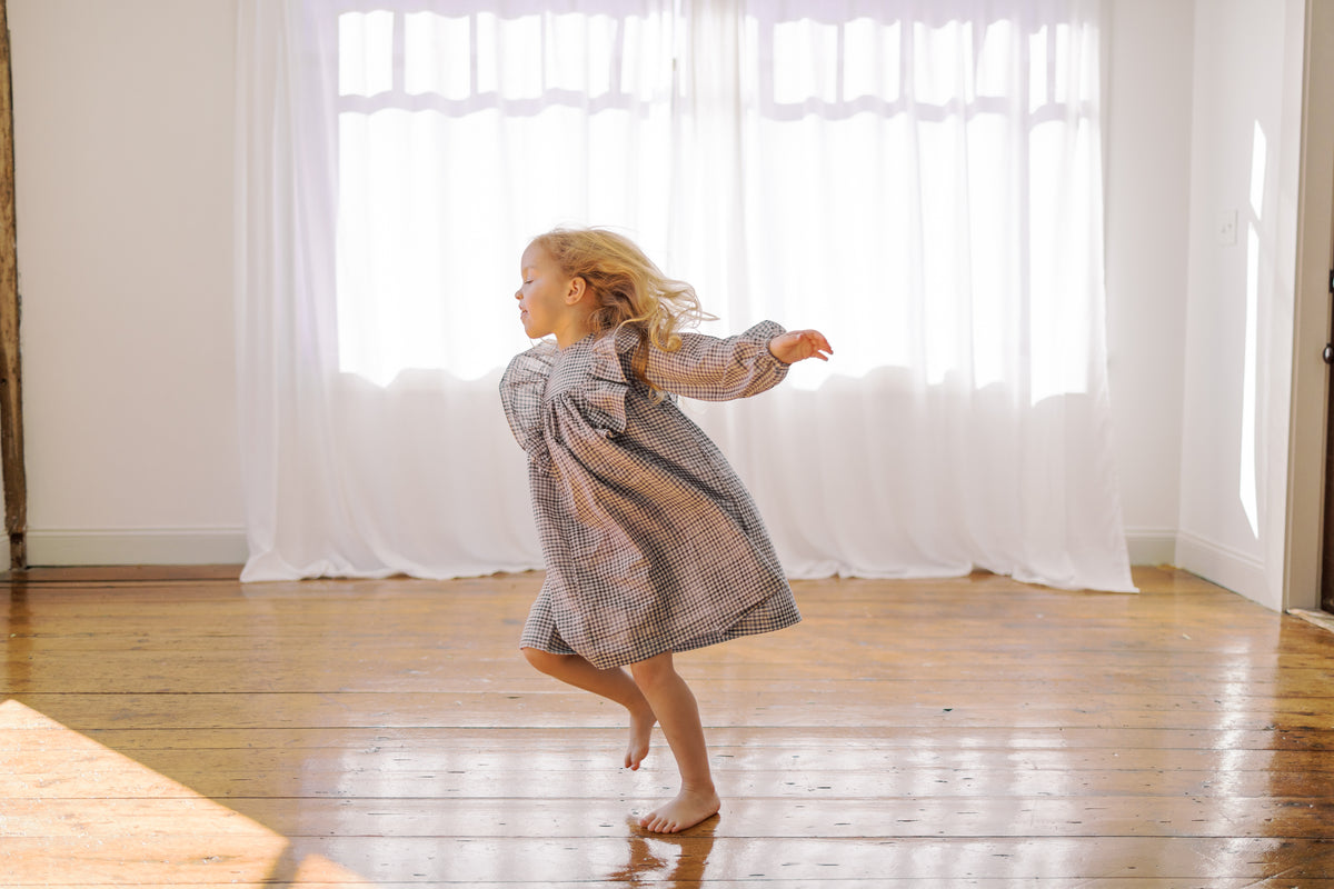 A young girl with blonde hair is barefoot, twirling in a loose, long-sleeved plaid dress or tunic in a bright room with white sheer curtains and a polished wooden floor.