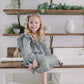 A young girl with blonde hair sits barefoot on a white wooden table, wearing a long-sleeve gingham dress in a checkered gray and white pattern. She is posed in front of a rustic wall with floating wooden shelves holding green plants and a white ceramic pitcher.