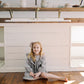 A young girl with blonde hair sits cross-legged on a wooden floor in front of a white kitchen island, wearing a purple and white check. The background features a white shiplap wall, wooden floating shelves with greenery and ceramic pitchers, and warm sunlight streaming across the floor 
