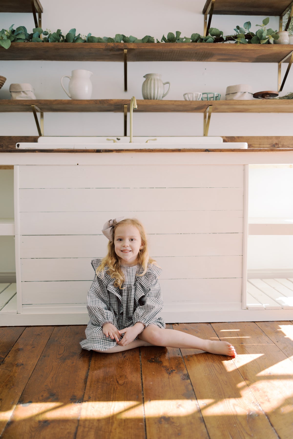 A young girl with blonde hair sits cross-legged on a wooden floor in front of a white kitchen island, wearing a purple and white check. The background features a white shiplap wall, wooden floating shelves with greenery and ceramic pitchers, and warm sunlight streaming across the floor 