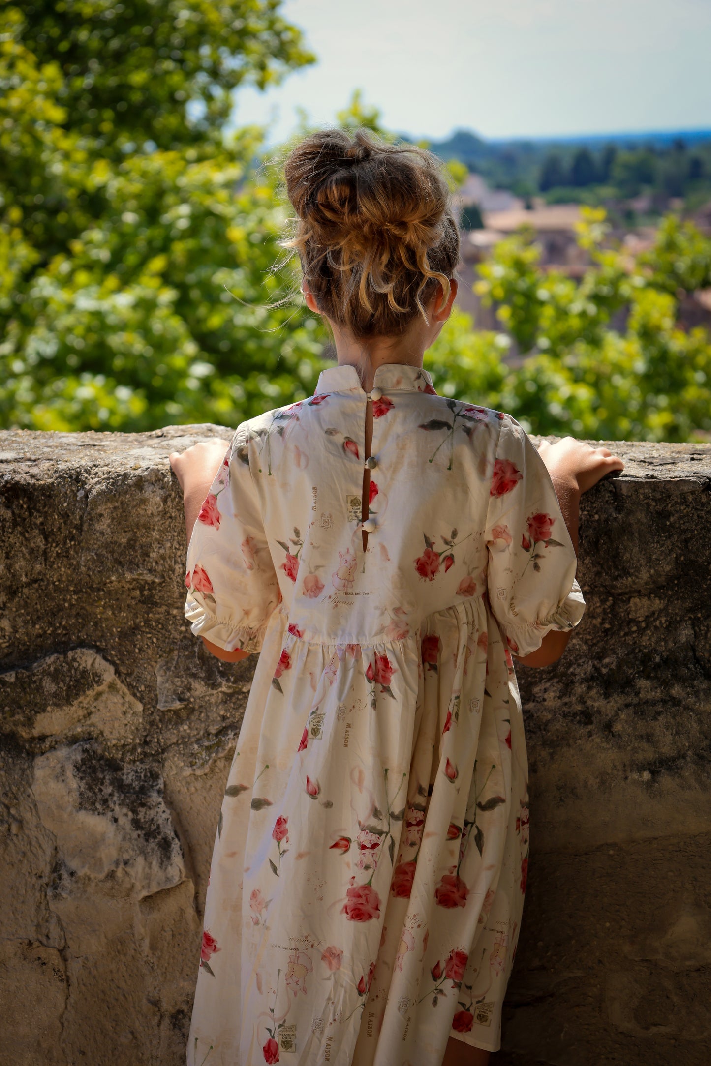Woman in a floral dress standing against a stone wall with greenery in the background