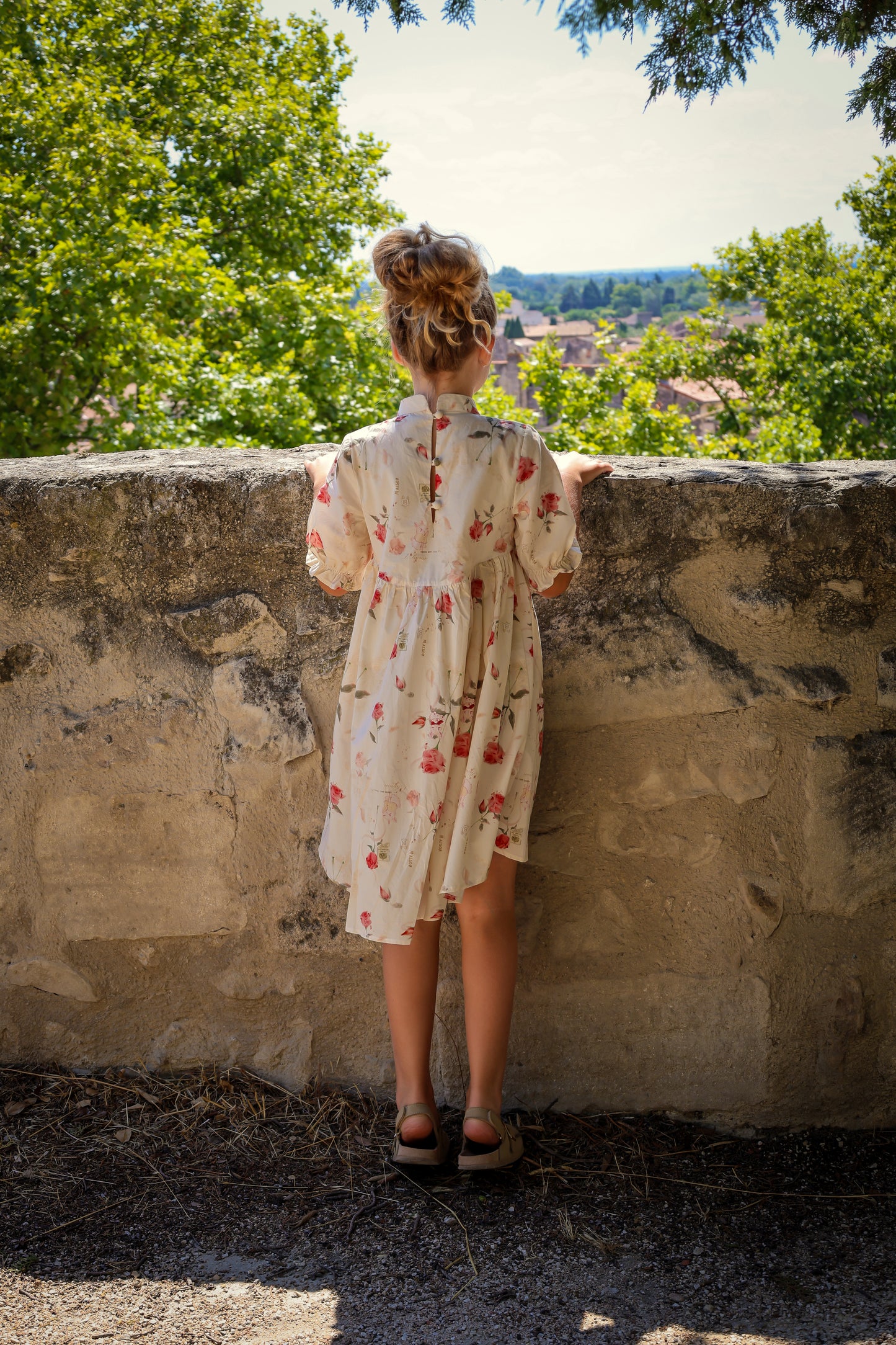 Woman in a floral dress standing against a stone wall with a scenic view in the background