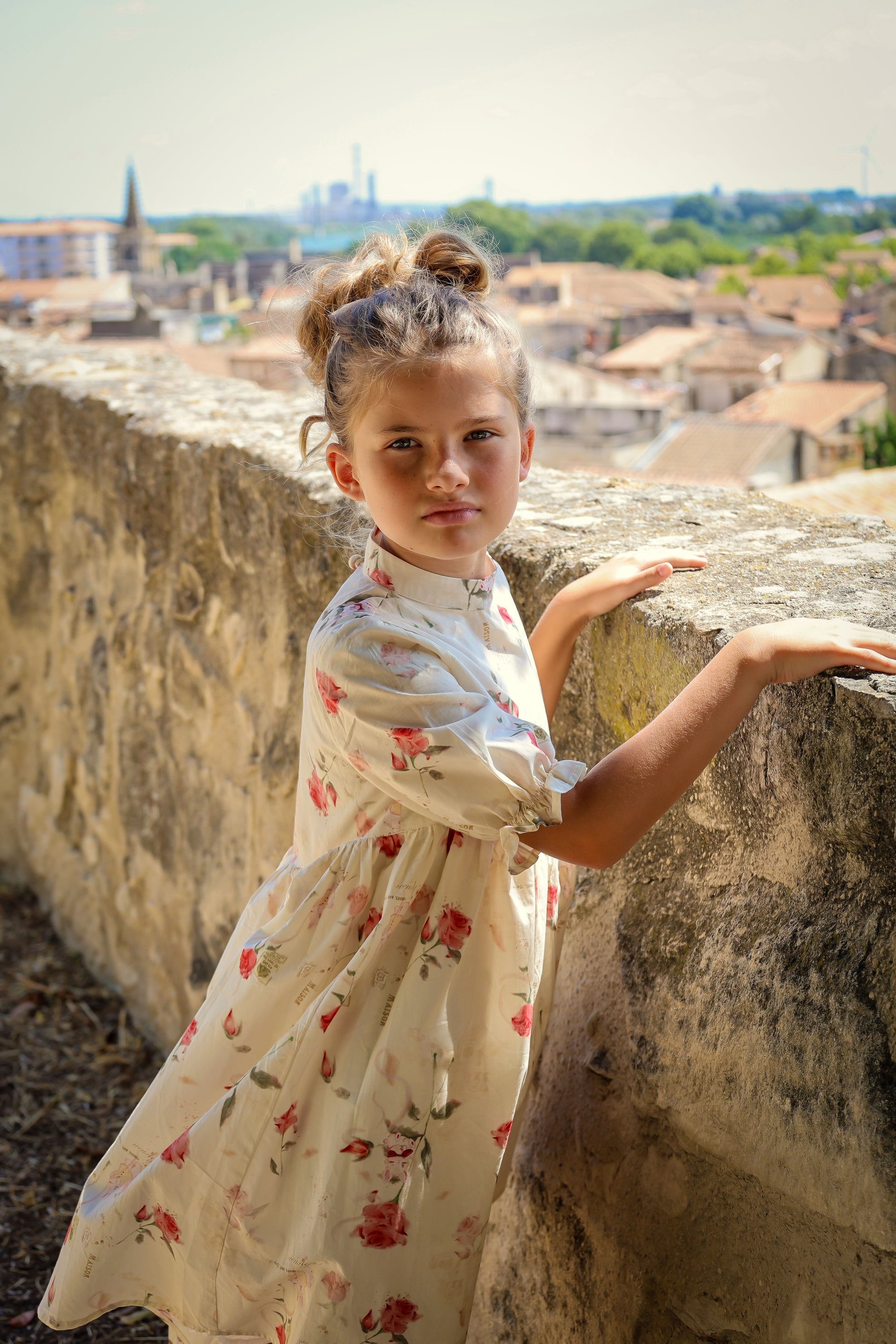 Young girl in a floral dress standing on a stone wall with a cityscape in the background