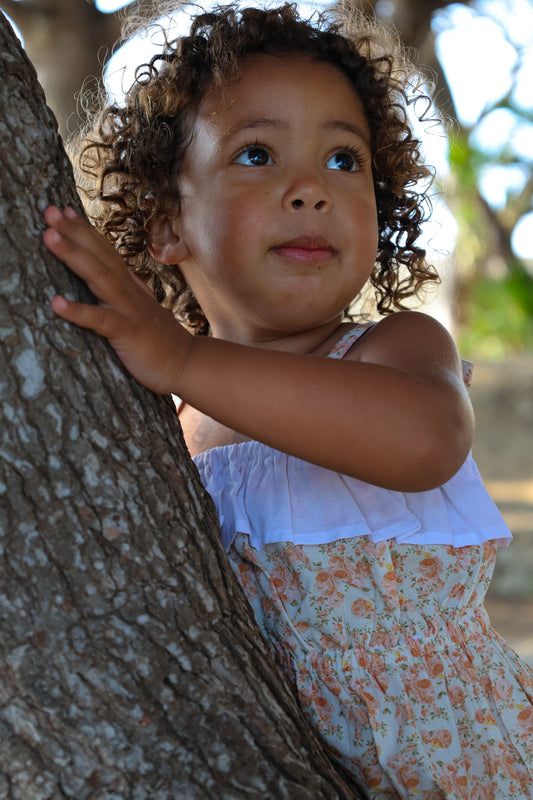 Child in a floral romper with adjustable straps peeking from behind a tree