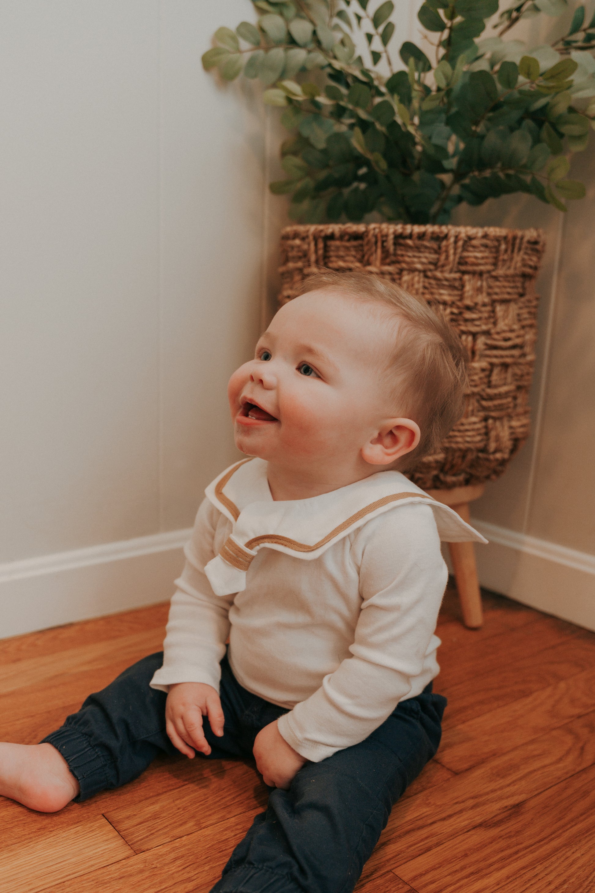 A baby wearing a white sailor-style collar bib with a knotted tie, sitting on the floor.
