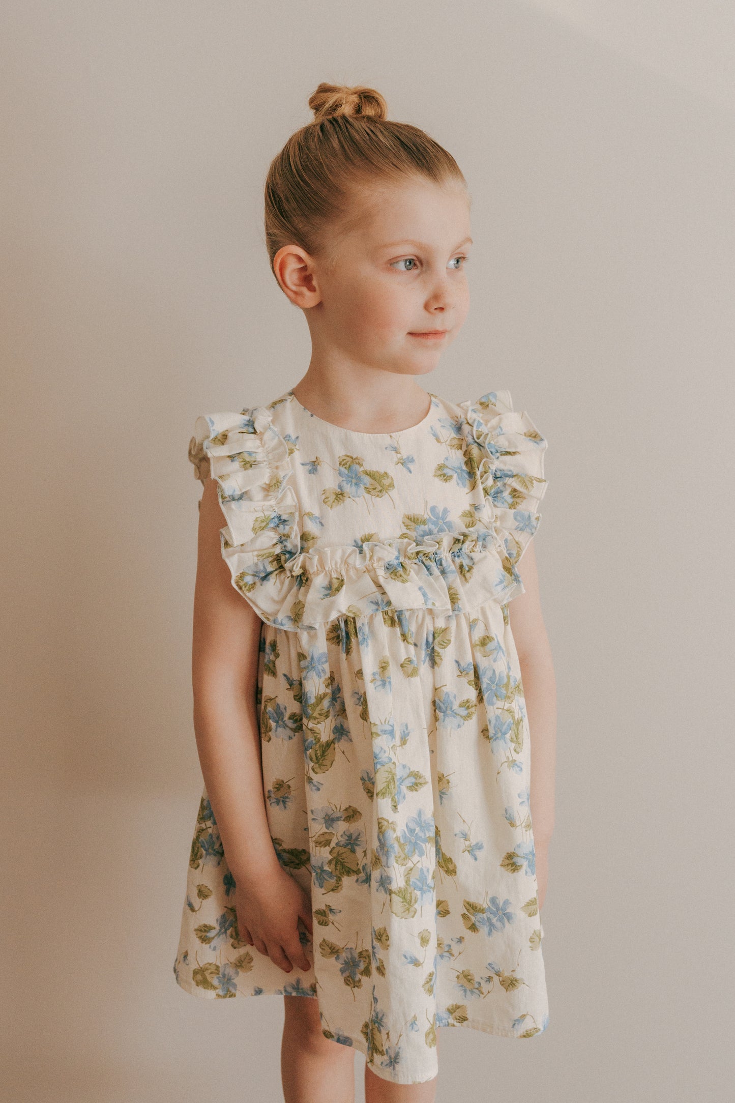 A young girl wearing a sleeveless, floral-print cotton dress with ruffled details.