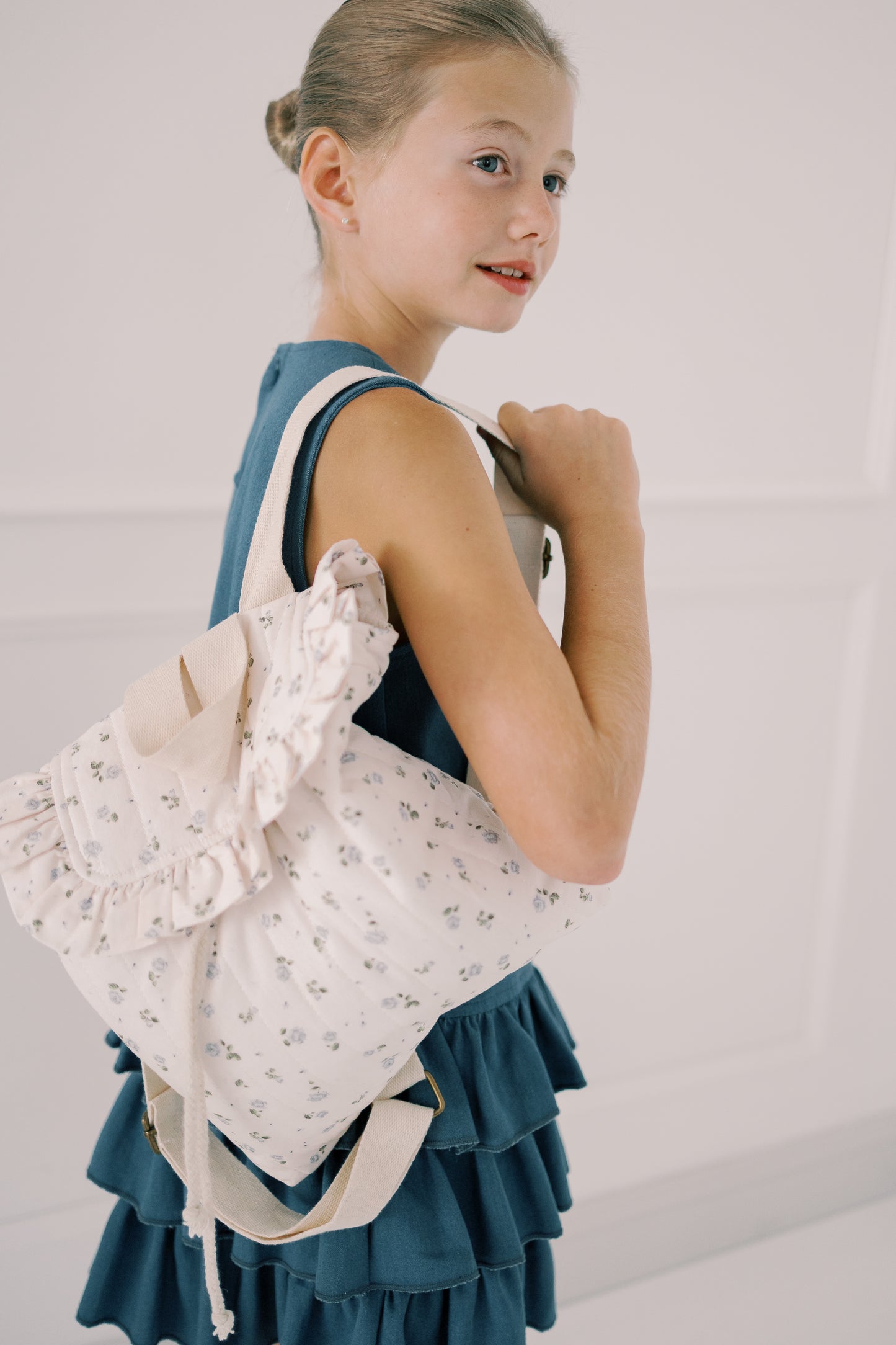 Young girl wearing a blue dress with a white floral backpack against a plain background