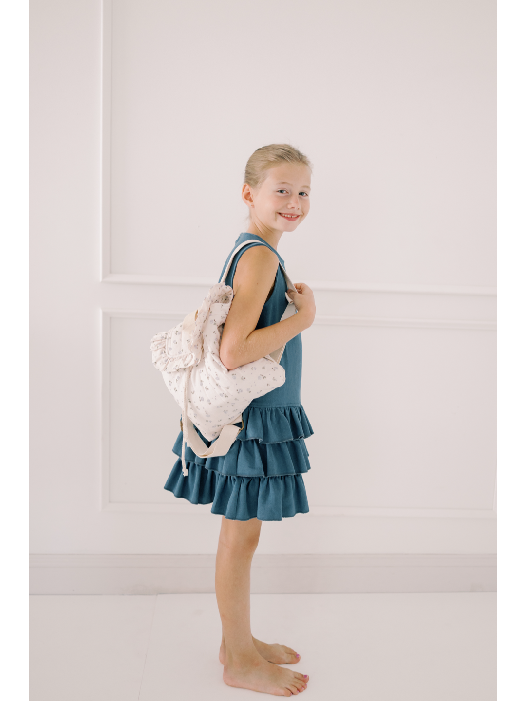 Young girl in a blue dress holding a white tote bag against a light-colored wall.