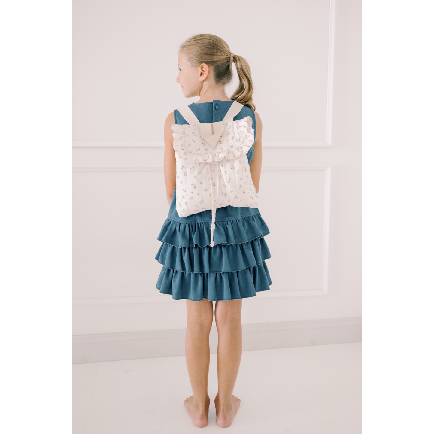 Girl wearing a denim dress with a white lace backpack on a plain background