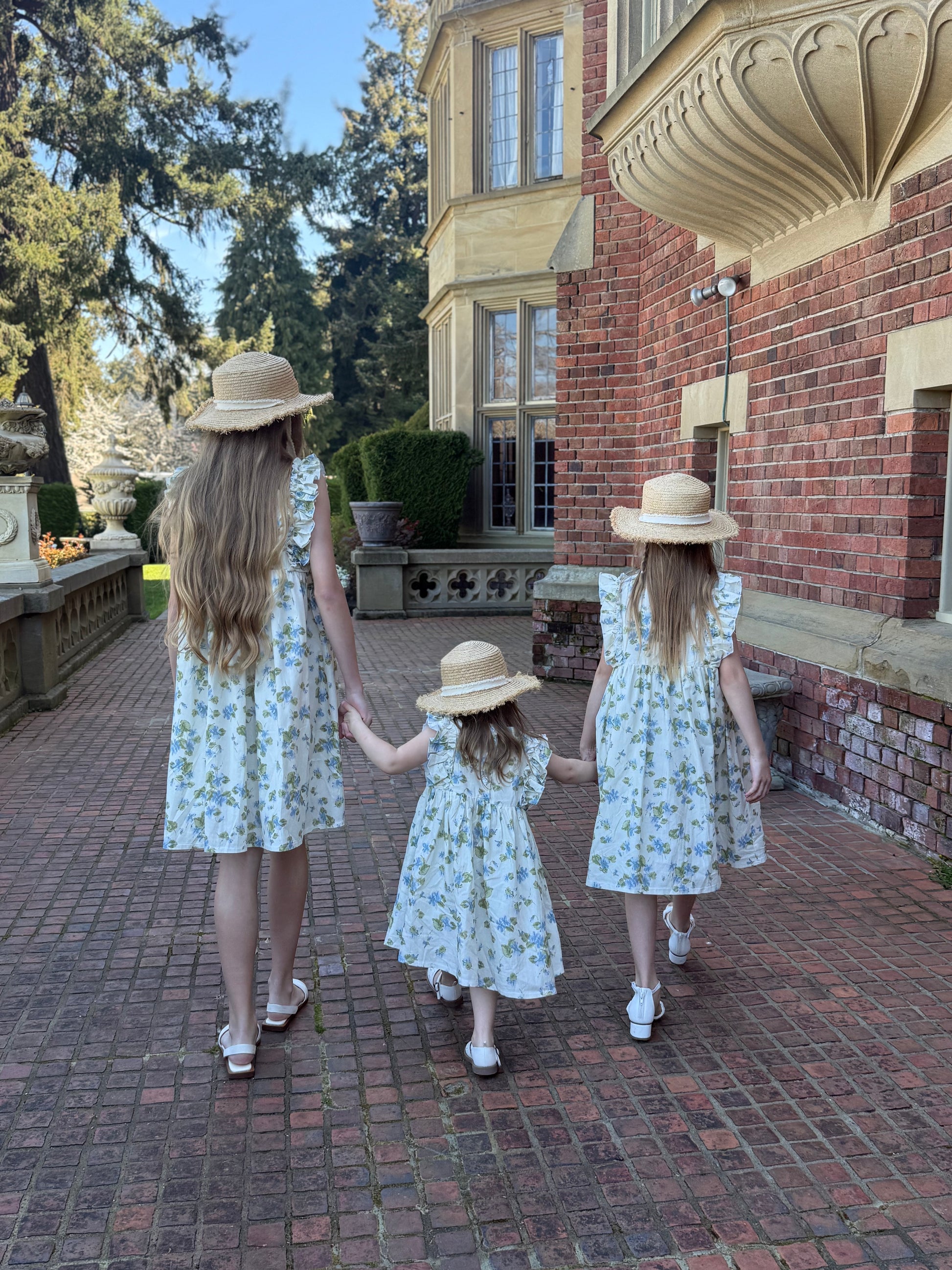 Three sisters in matching dresses and hats walking together on a brick path in front of a brick building.