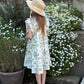 Young girl in a floral dress and sun hat standing in a garden with white flowers.