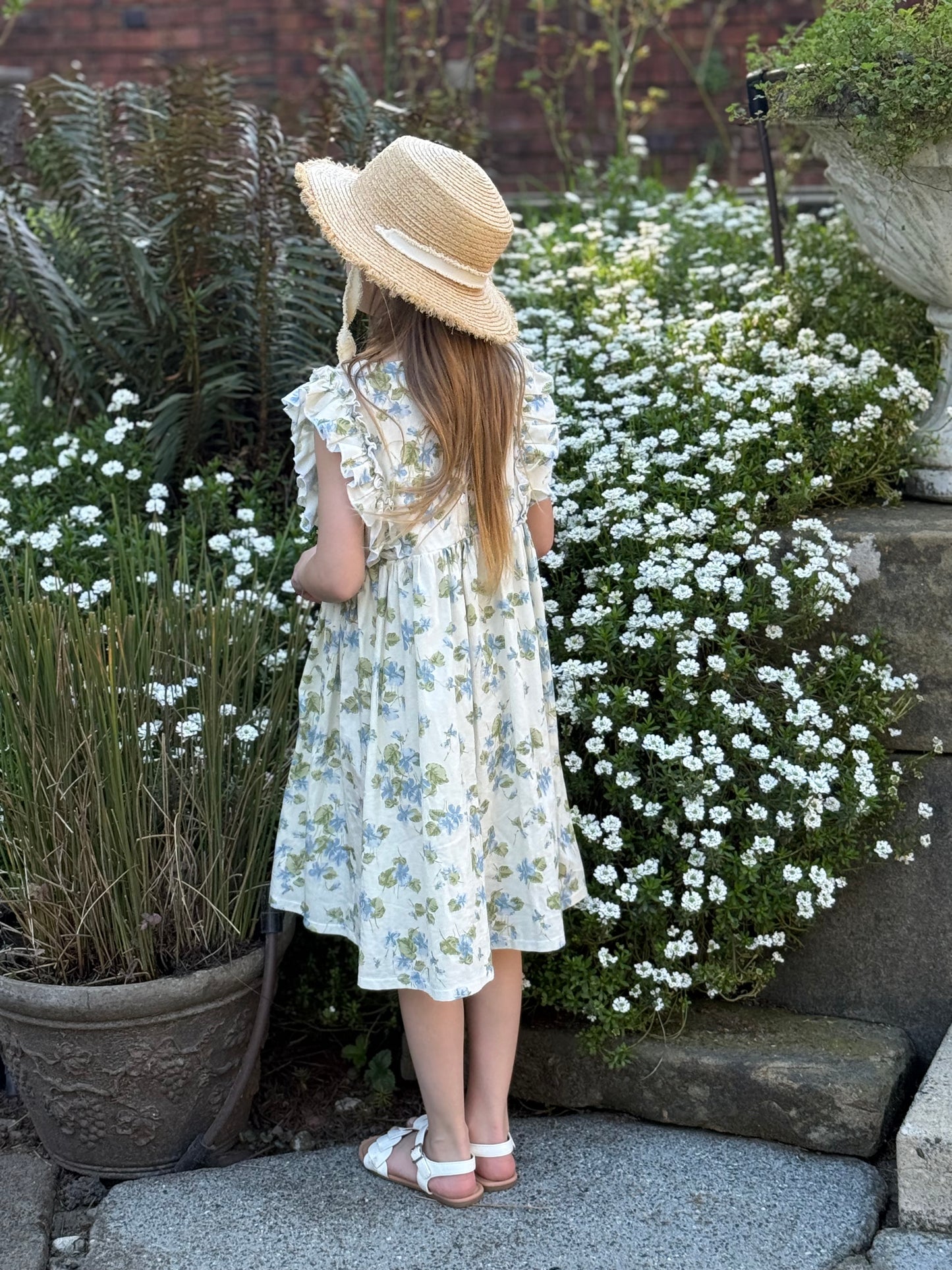 Young girl in a floral dress and sun hat standing in a garden with white flowers.