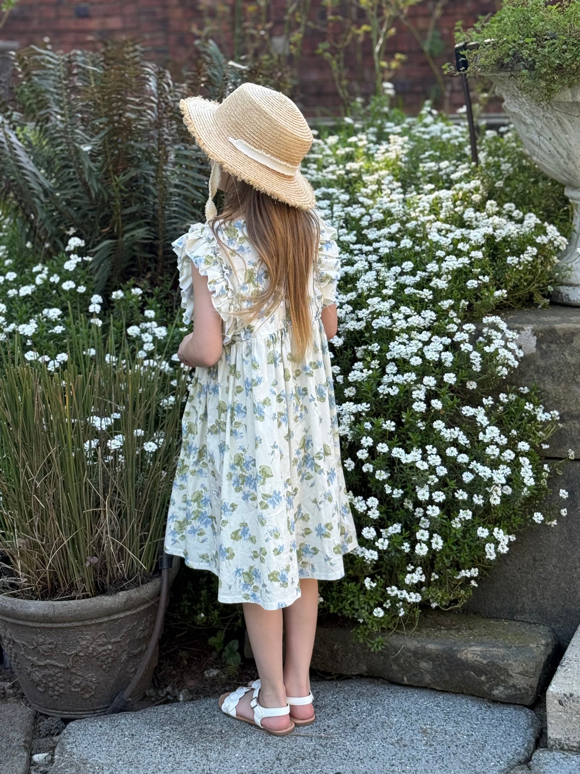 Young girl in a floral dress and sun hat standing in a garden with white flowers.