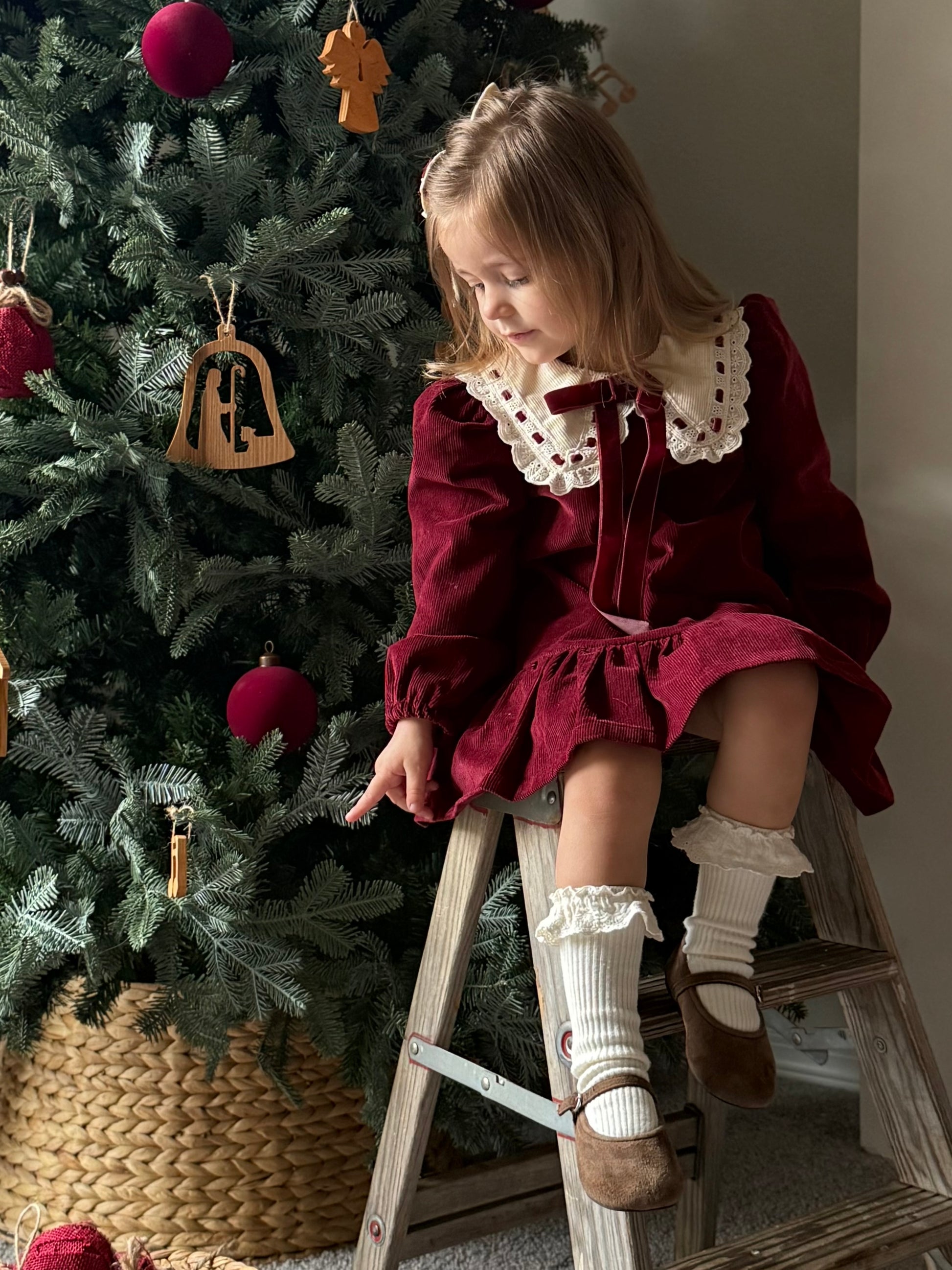 Child in a red dress sitting on a ladder next to a decorated Christmas tree.