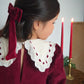Child wearing a burgundy dress with a white lace collar, standing in front of candles and a Christmas tree.