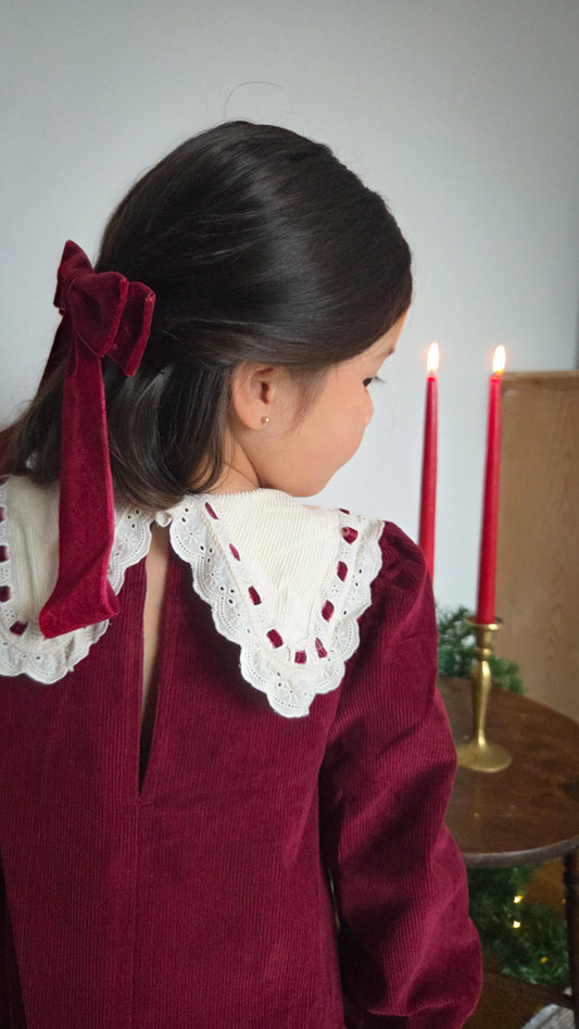 Child wearing a burgundy dress with a white lace collar, standing in front of candles and a Christmas tree.