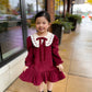 Young girl in a burgundy dress with white lace collar standing on a sidewalk.