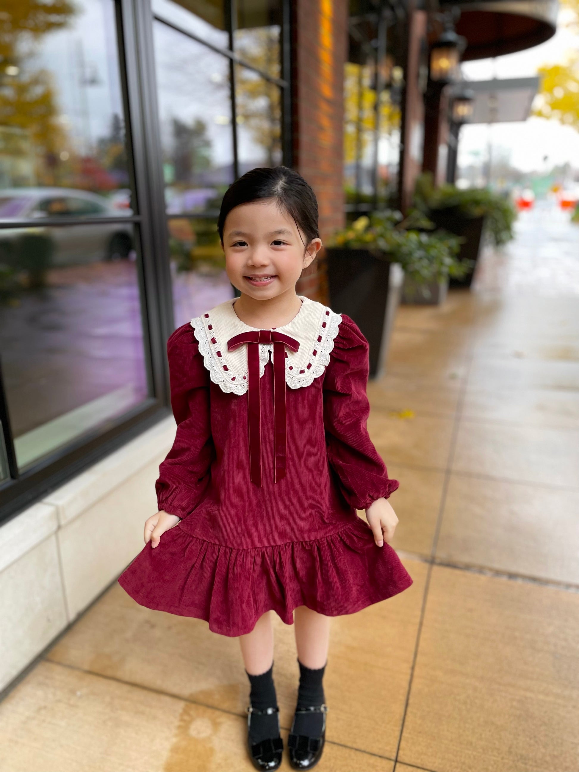 Young girl in a burgundy dress with white lace collar standing on a sidewalk.