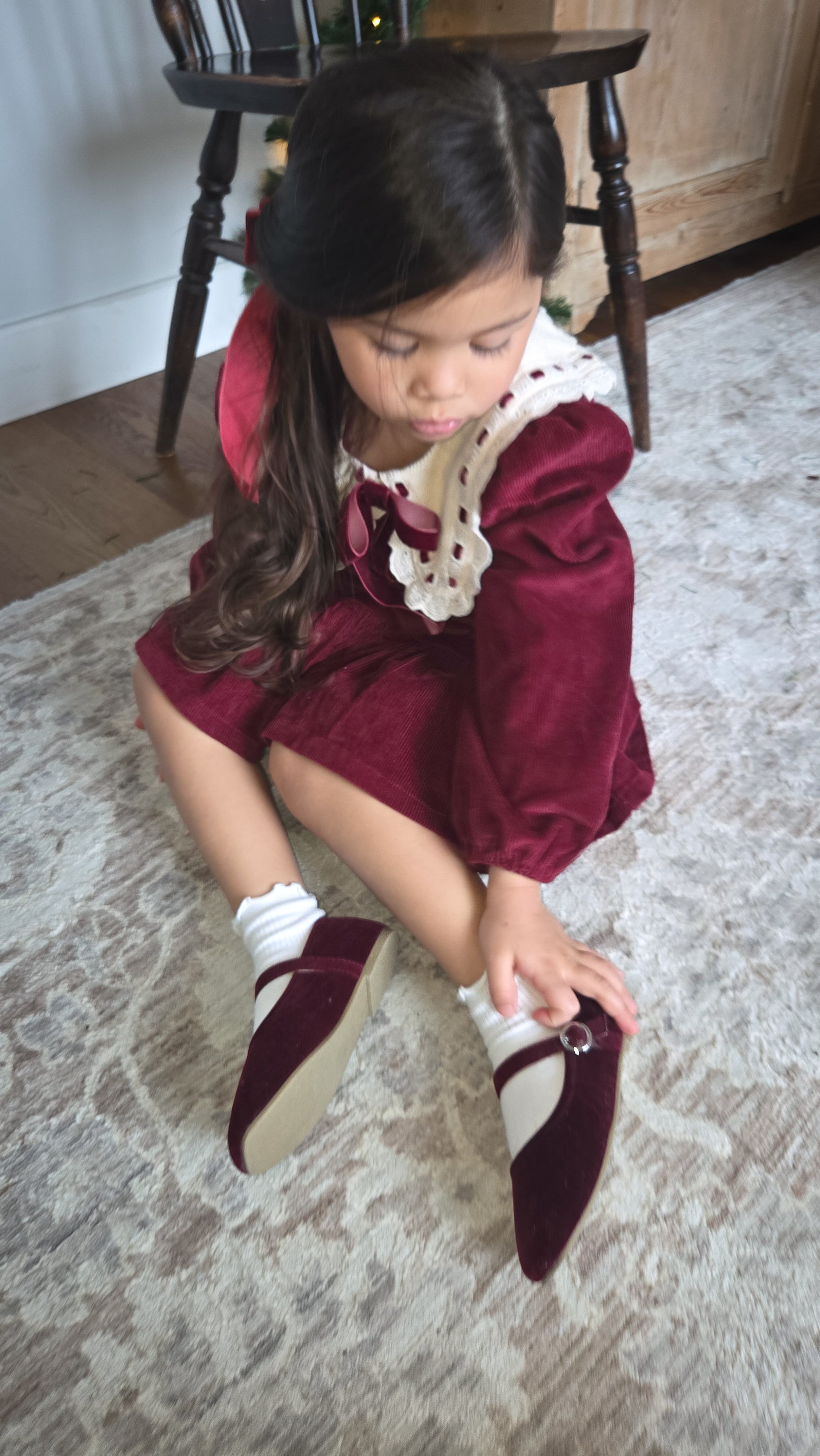 Young girl in a burgundy dress sitting on a patterned rug.