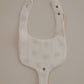 A floral pacifier bib with a neutral beige background and small flowers printed across it. The bib has snap closures and is designed to hold a pacifier.
