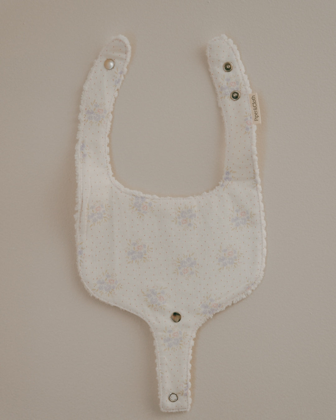 A floral pacifier bib with a neutral beige background and small flowers printed across it. The bib has snap closures and is designed to hold a pacifier.