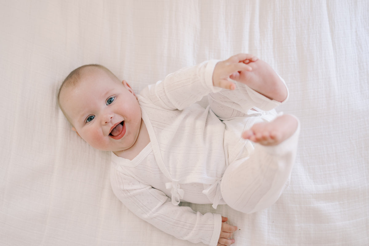 A smiling baby lying on a white textured blanket, wearing a white long-sleeve ribbed romper.