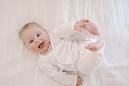 A smiling baby lying on a white textured blanket, wearing a white long-sleeve ribbed romper.