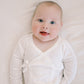 A smiling baby lying on a white textured blanket, wearing a white long-sleeve wrap bodysuit with a small bow detail on the chest.