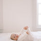 A baby lying on a white bed, wearing a white long-sleeve knit romper and looking toward the camera in a bright, minimalist room.