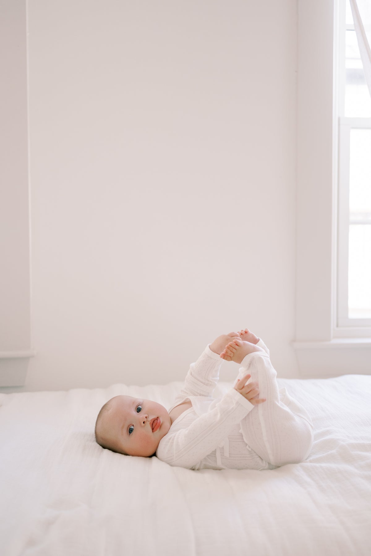 A baby lying on a white bed, wearing a white long-sleeve knit romper and looking toward the camera in a bright, minimalist room.