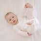 A baby lying on a white textured blanket, wearing a white long-sleeve ribbed bodysuit and looking up with one hand while holding up the other leg.