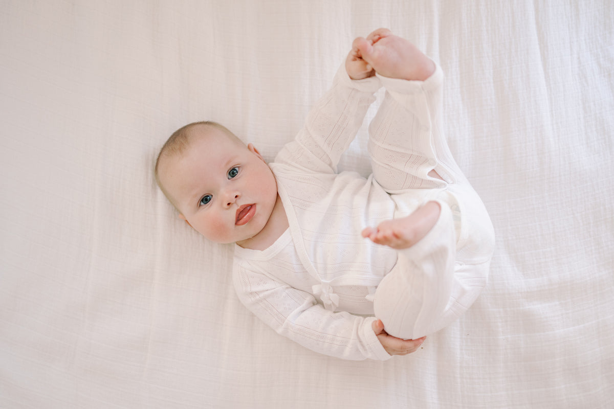 A baby lying on a white textured blanket, wearing a white long-sleeve ribbed bodysuit and looking up with one hand while holding up the other leg.