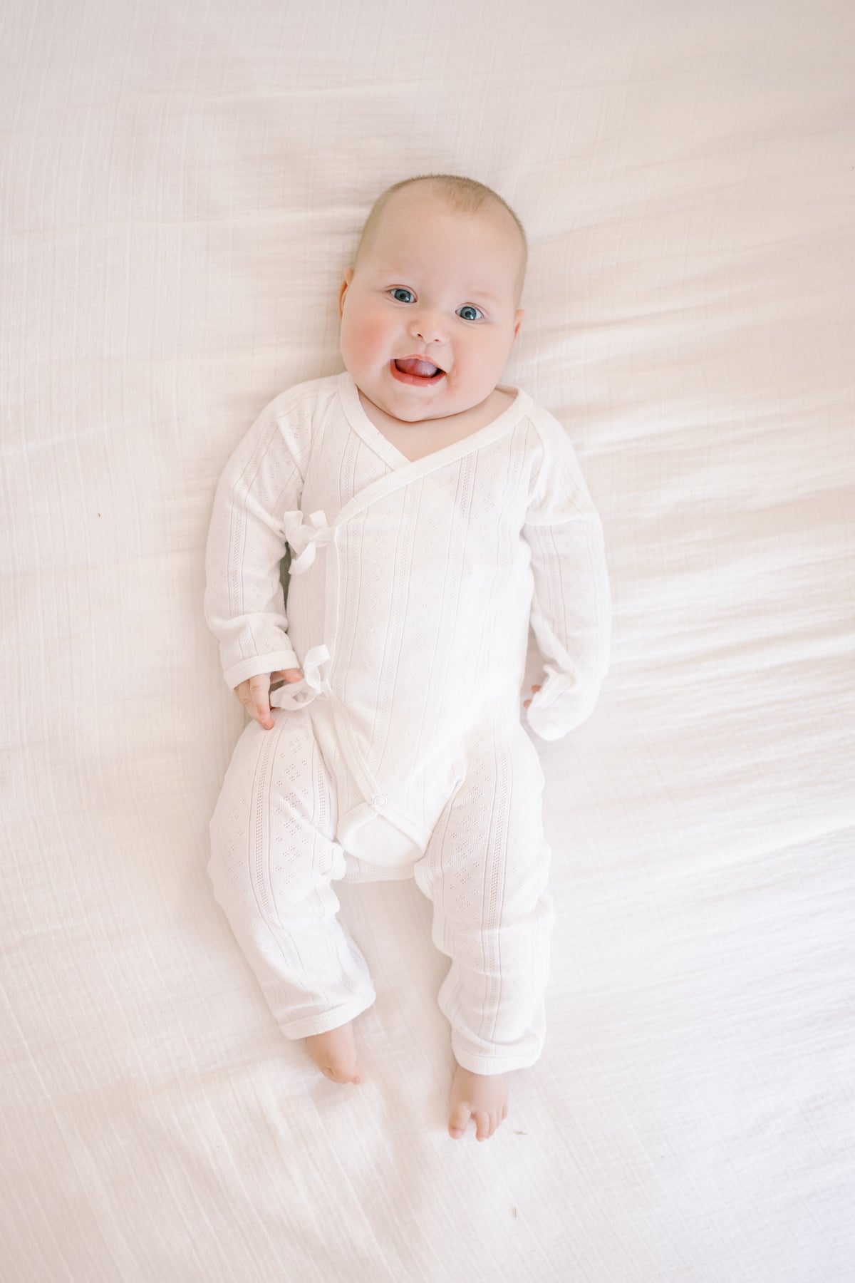 A baby lying on a white textured blanket, wearing a white ribbed long-sleeve footless bodysuit with side ties.