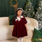 Young girl in a burgundy dress with white collar standing in a festive room with Christmas trees and decorations.
